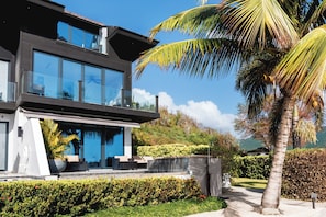 A modern two-story house with large glass windows, a balcony, and a well-maintained garden with palm trees and hedges. - Beach House 3: Luxury 3-Bedroom Beach House with Private Pool, Antigua (Urlings)