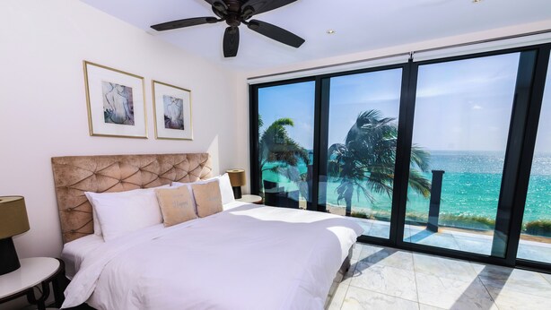 Ein Schlafzimmer mit einem großen Bett, einem Deckenventilator und Blick auf den Ozean durch raumhohe Fenster.