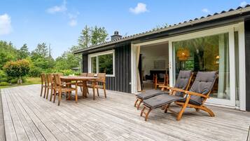 En træterrasse med et spisebord og stole, en liggestol og en glasdør, der fører ind i et rum.