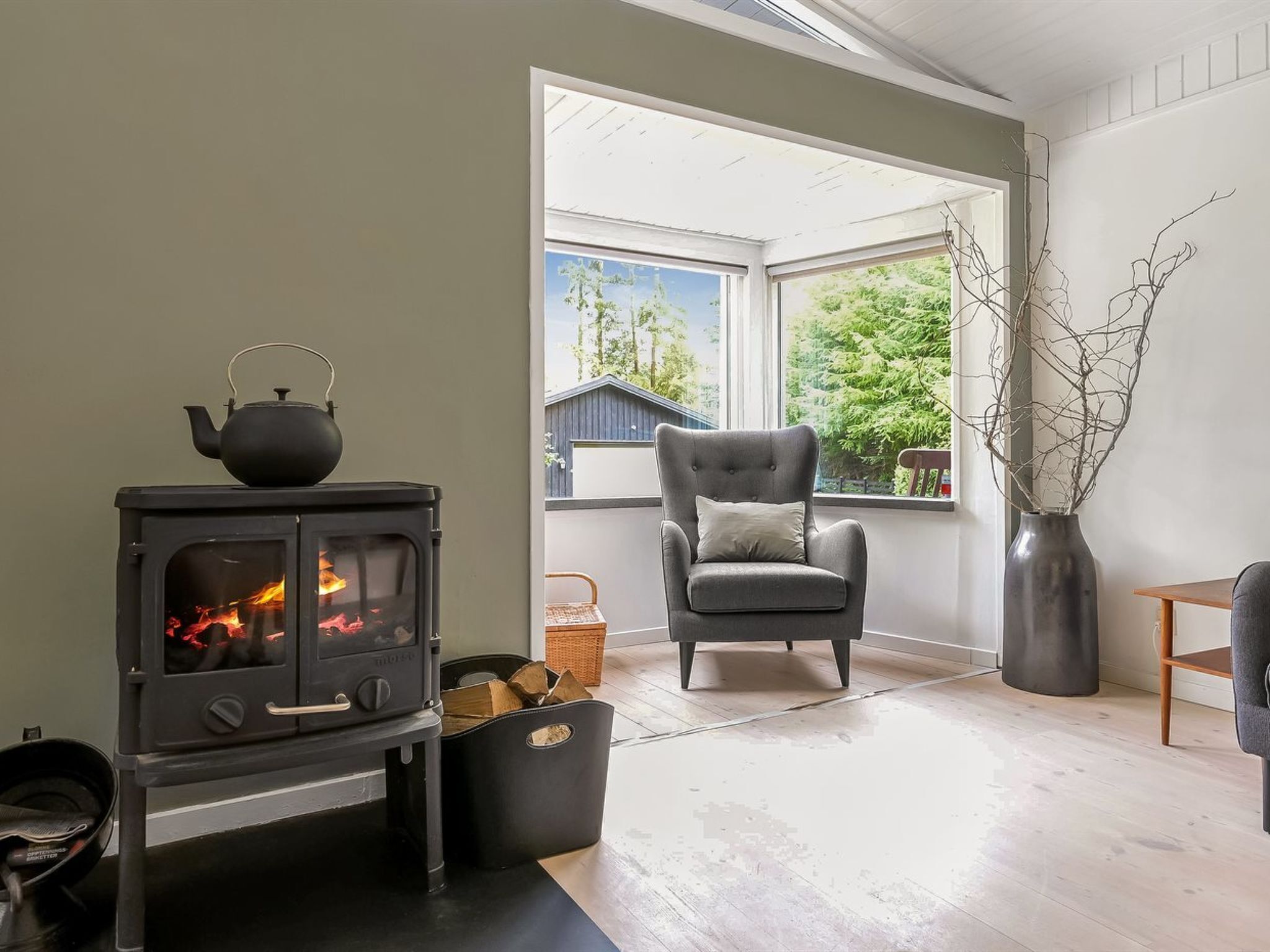 A cozy room with a wood-burning stove, a comfortable armchair, and a large window offering a view of trees and a building outside.
