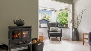 A cozy room with a wood-burning stove, a comfortable armchair, and a large window offering a view of trees and a building outside.