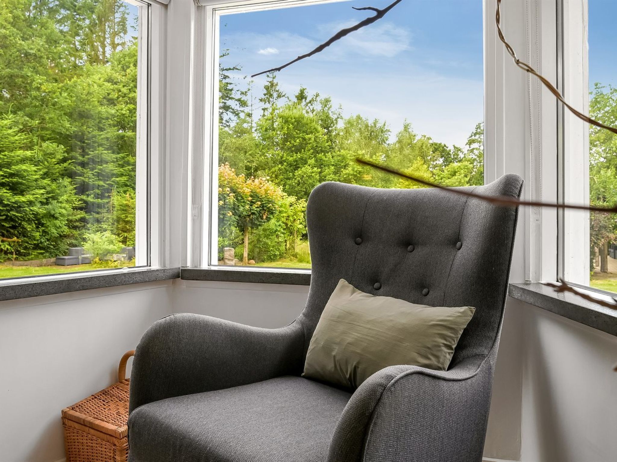 Un fauteuil gris, recouvert d’un tissu d’ameublement, doté d’un dossier capitonné et d’un coussin, placé dans un coin et offrant une vue sur un paysage verdoyant grâce à une grande fenêtre.