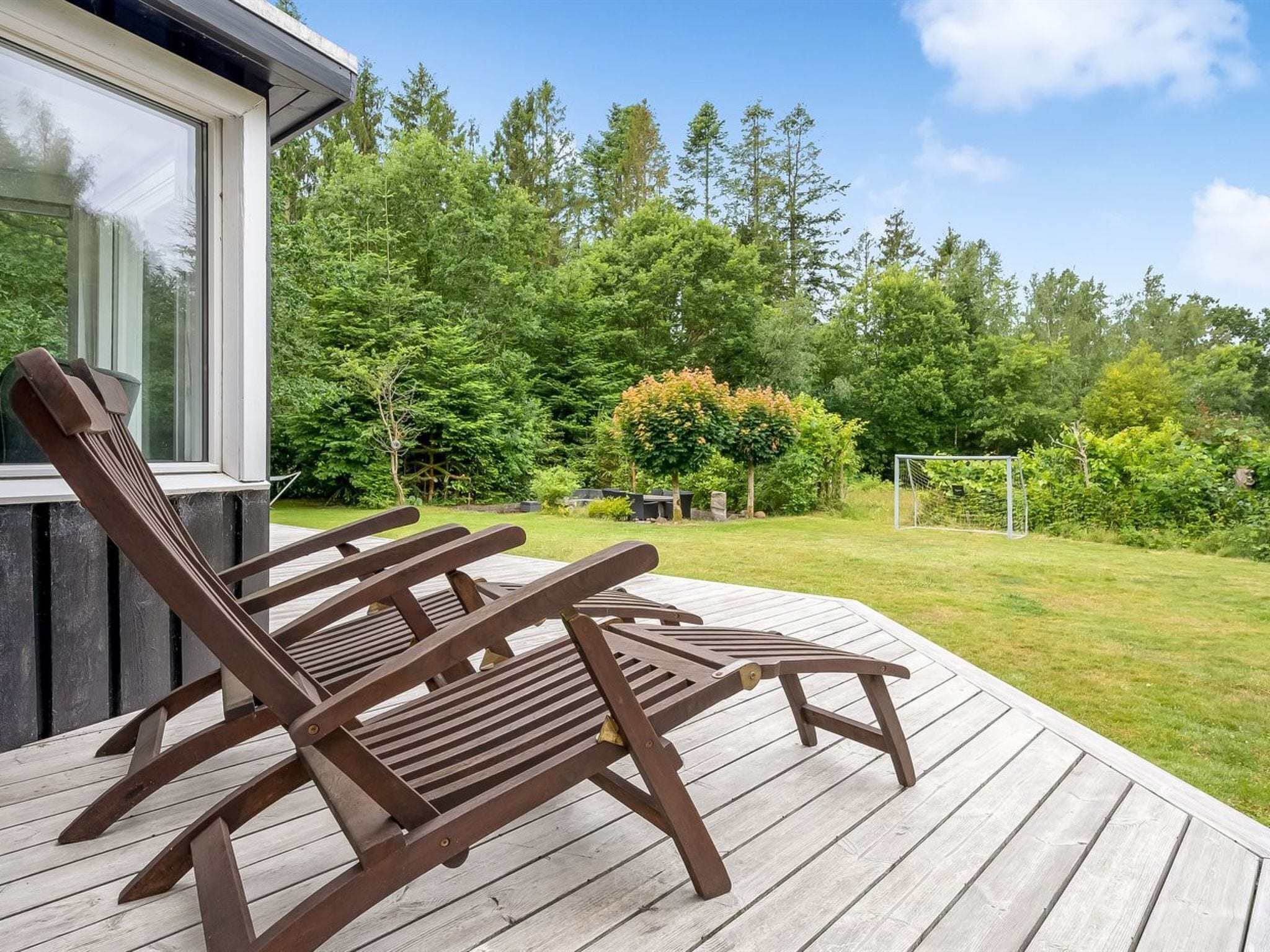A wooden deck with two lounge chairs, a house with a large window, and a lush green garden with trees and a soccer goal.