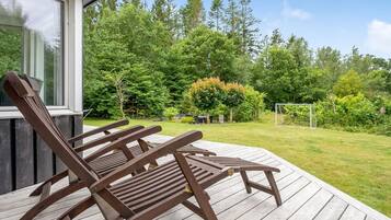 A wooden deck with two lounge chairs, a house with a large window, and a lush green garden with trees and a soccer goal.