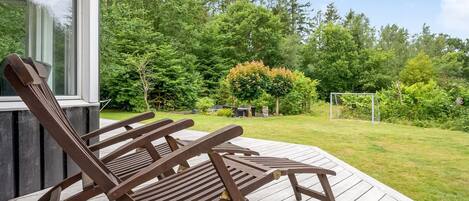 A wooden deck with two lounge chairs, a house with a large window, and a lush green garden with trees and a soccer goal.