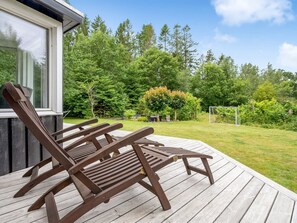 Une terrasse en bois avec deux chaises longues, une maison dotée d’une grande fenêtre et un jardin luxuriant, verdoyant, avec des arbres et un but de football.