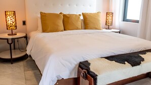 A neatly made bed with white linens and a brown headboard, flanked by two bedside tables with lamps, and a window with curtains. - Beach House 5: Luxury 3-Bedroom Beach House with Private Pool, Antigua (Urlings)