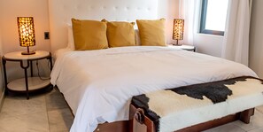 A neatly made bed with white linens and a brown headboard, flanked by two bedside tables with lamps, and a window with curtains. - Beach House 5: Luxury 3-Bedroom Beach House with Private Pool, Antigua (Urlings)