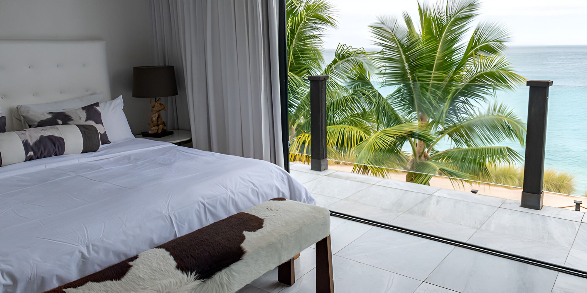 A bedroom with a large bed, a nightstand, a lamp, and a view of the ocean and palm trees.