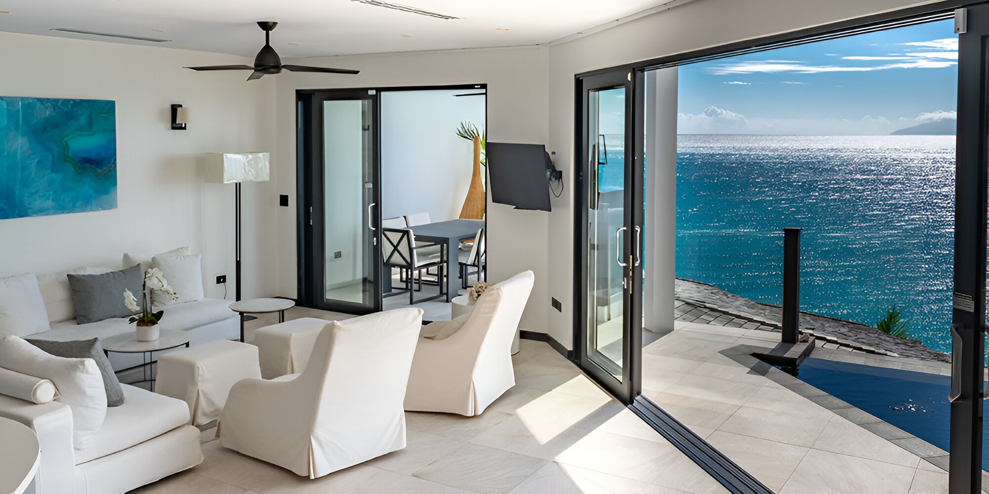 A modern living room with a sofa, chairs, and a dining table. There is a large window with a view of the ocean.