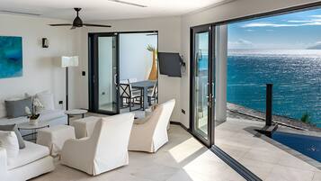 A modern living room with a sofa, chairs, and a dining table. There is a large window with a view of the ocean.