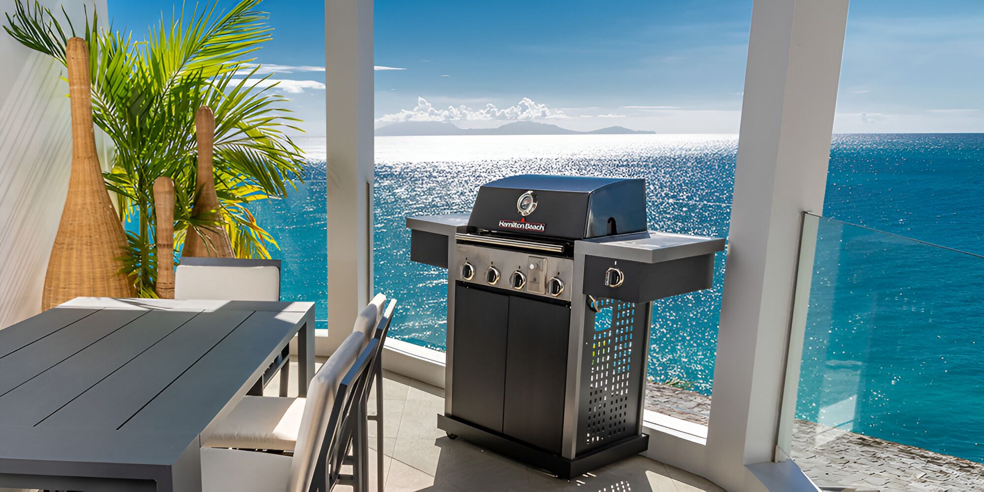 An outdoor grill with a view of the ocean, a table, chairs, and a potted plant.