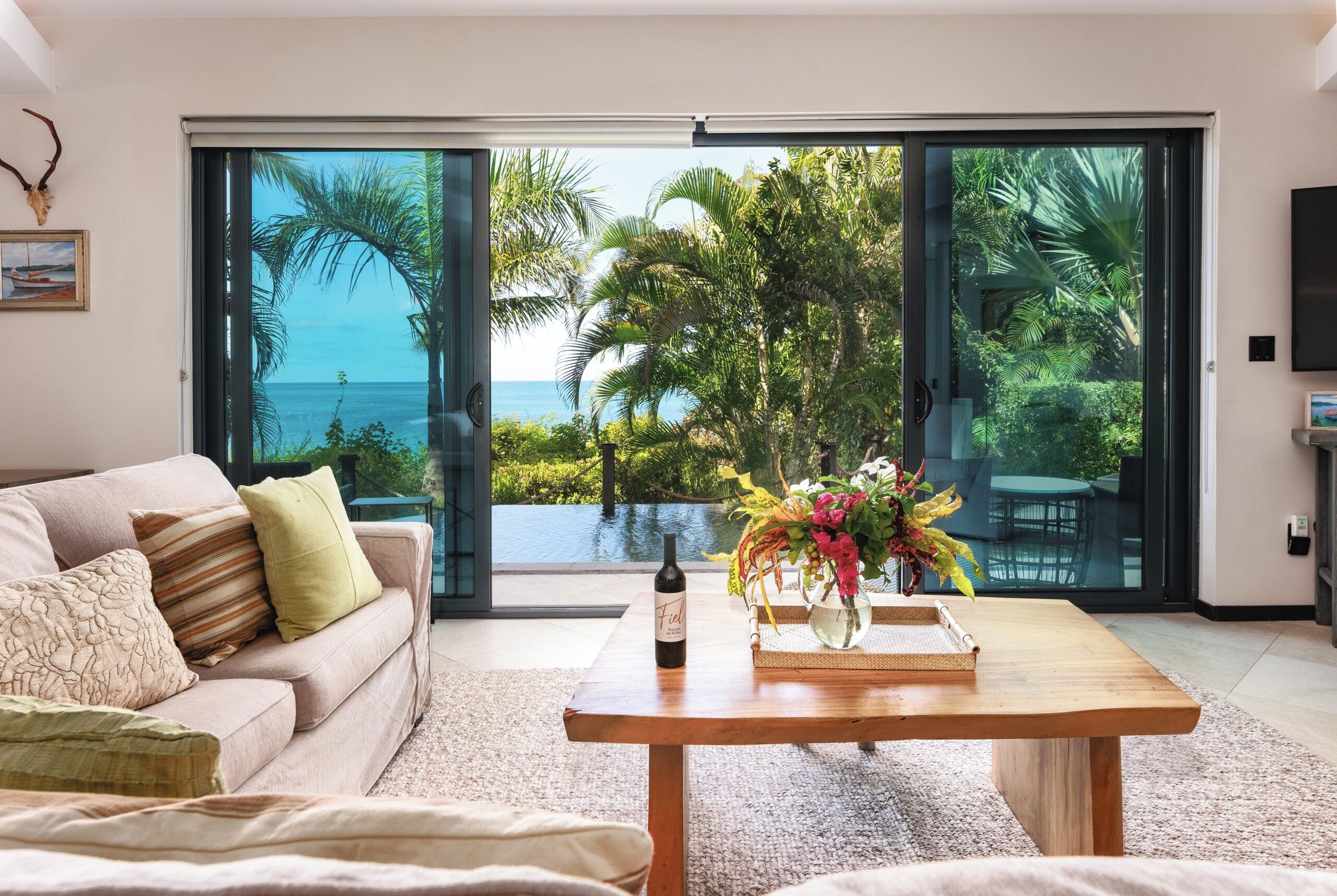 A modern living room with a large sliding glass door leading to a pool and ocean view, a sofa with cushions, a wooden coffee table with a bottle of wine and a vase of flowers.