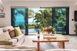 A modern living room with a large sliding glass door leading to a pool and ocean view, a sofa with cushions, a wooden coffee table with a bottle of wine and a vase of flowers. - Oasis: Luxury 2-Bedroom Villa with Boardwalk to Darkwood & Ffryes Beach, Antigua (Urlings)