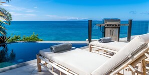 Zona de descanso junto a la piscina con dos tumbonas y una parrilla, con vistas al mar azul y transparente.
