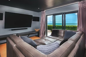 A modern living room with a large flat-screen TV, leather sofas, and a view of the ocean. - Spacious 4BR Estate with Private Gym & Outdoor Living (Urlings)