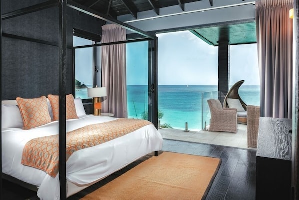 Un dormitorio moderno con una cama grande, vistas al océano y una zona de estar con una silla y una mesa.