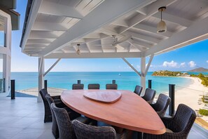 Un área de comedor al aire libre con sillas de mimbre y una mesa de madera, con vistas a una playa y al mar.