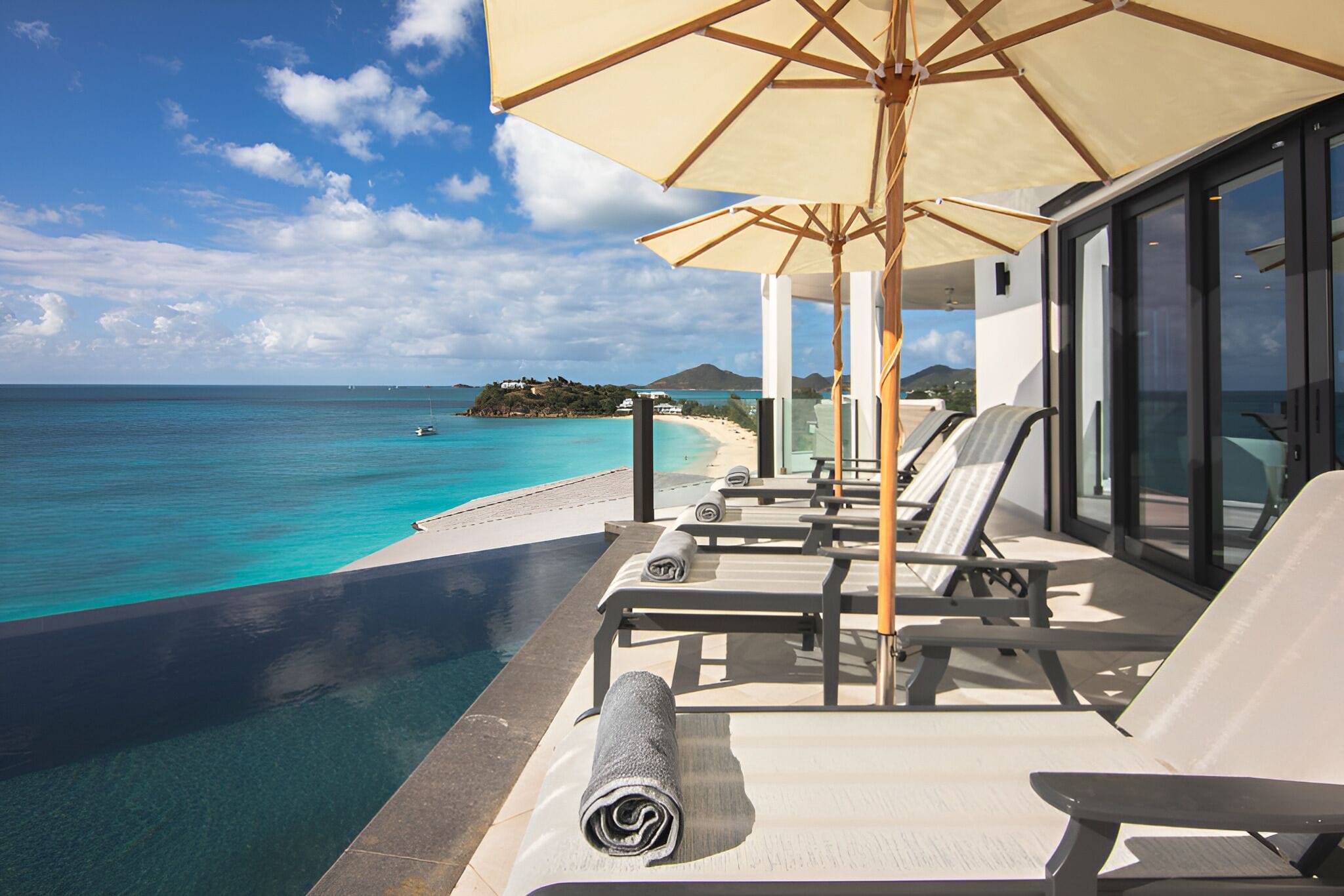 A poolside area with lounge chairs and a large umbrella, overlooking a clear blue sea and a distant island.