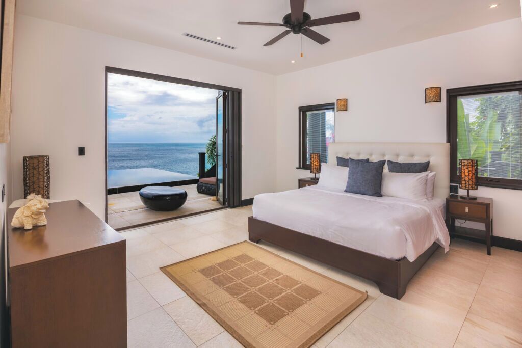 Ein Schlafzimmer mit einem großen Bett, einem Deckenventilator, einem Schreibtisch und Blick auf den Ozean.