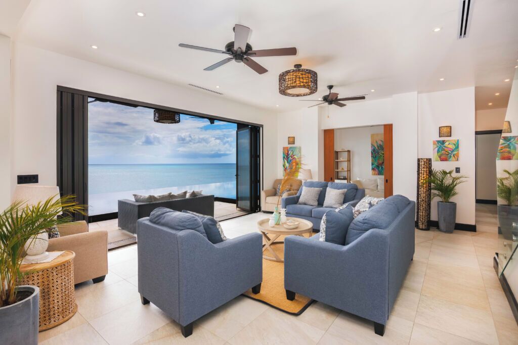 Ein geräumiges Wohnzimmer mit einem großen Schiebeglasfenster, das auf einen Balkon mit Meerblick führt. Zwei blaue Sofas, ein Couchtisch und ein Deckenventilator sind vorhanden.