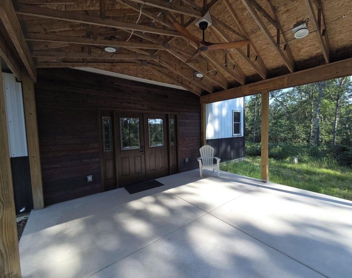 A covered porch with a wooden ceiling and a concrete floor, a white chair, and a view of a house through large windows.