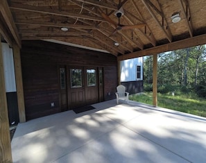 A covered porch with a wooden ceiling and a concrete floor, a white chair, and a view of a house through large windows. - Lovely 3-bedroom newly constructed barndominium perfect for relaxing stays. (Baldwin)
