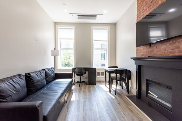 A modern living room with a black leather sofa, a dining table with chairs, a fireplace, and large windows.