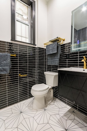 A modern bathroom with black and white tiled walls, a white toilet, a gold faucet, and a window.