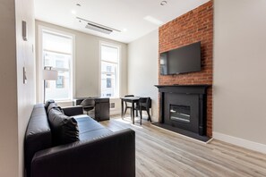 A modern living room with a sofa, a fireplace, a TV mounted on a brick wall, and large windows. - Modern 1-Bedroom Retreat with Fireplace in Downtown Brooklyn (Brooklyn)