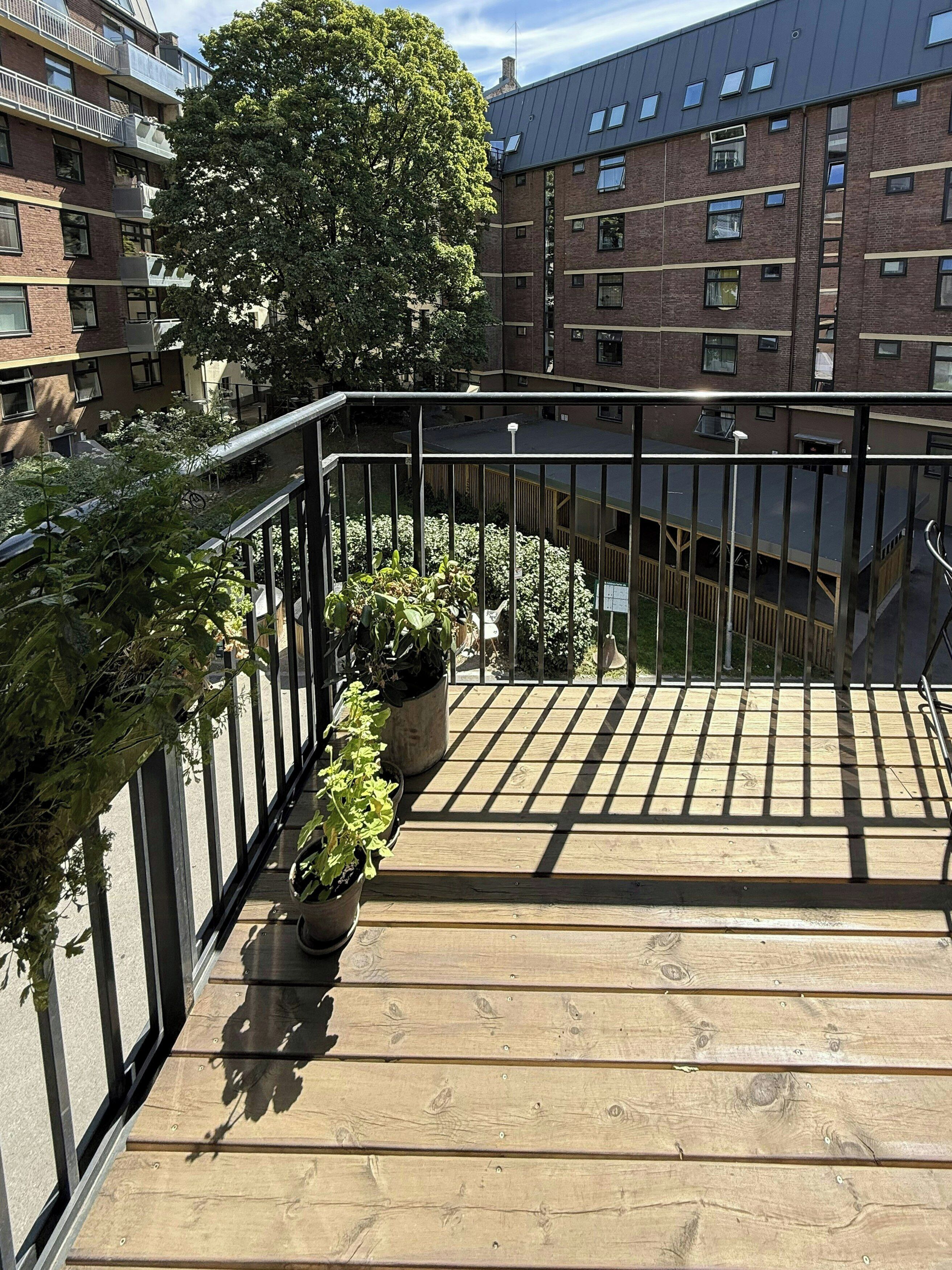 Una terraza de madera con plantas en macetas, una barandilla de metal y vistas a un edificio de ladrillo con varias ventanas.