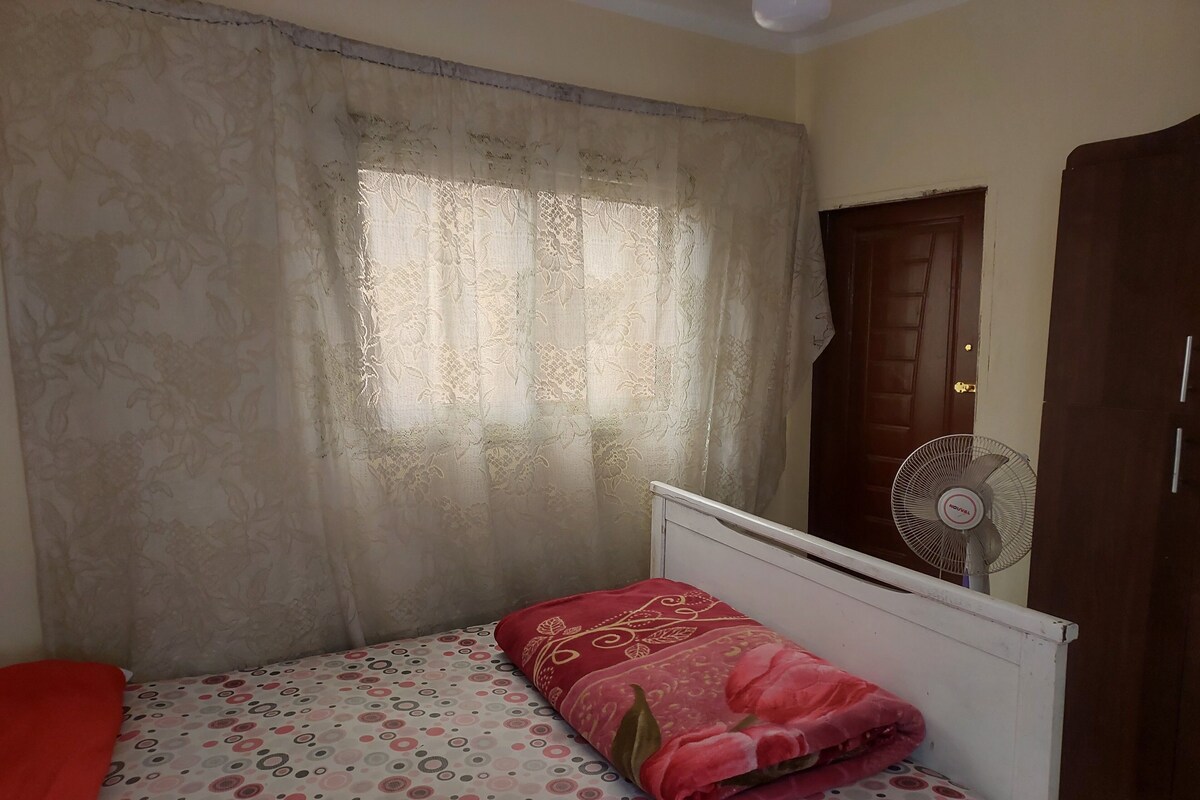 Une chambre à coucher avec un lit, un oreiller rouge, une fenêtre avec des rideaux, une porte en bois et un ventilateur sur pied.
