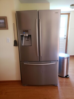 A stainless steel Samsung refrigerator with a water dispenser, positioned in a room with wooden flooring and a door in the background.