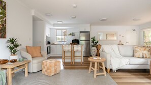 A modern living room with a sofa, a coffee table, and a dining area with a kitchen in the background. - Acacia Mollymook Beach | Pool & Tennis Court (Mollymook Beach)