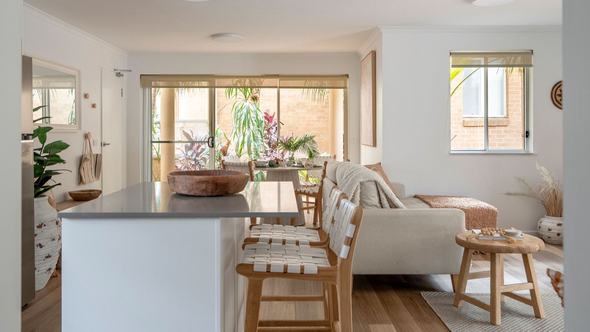 A modern kitchen with a central island, wooden chairs, a dining table, and a sofa with a view of the garden.