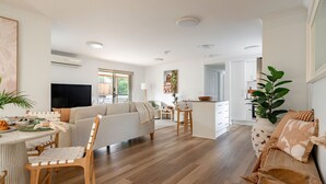 A spacious living area with a sofa, dining table, and chairs, a kitchen area with a dining table, and a large window providing natural light. - Acacia Mollymook Beach | Pool & Tennis Court (Mollymook Beach)