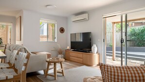 A modern living room with a flat-screen TV, a sofa, a coffee table, and a dining area with chairs. - Acacia Mollymook Beach | Pool & Tennis Court (Mollymook Beach)