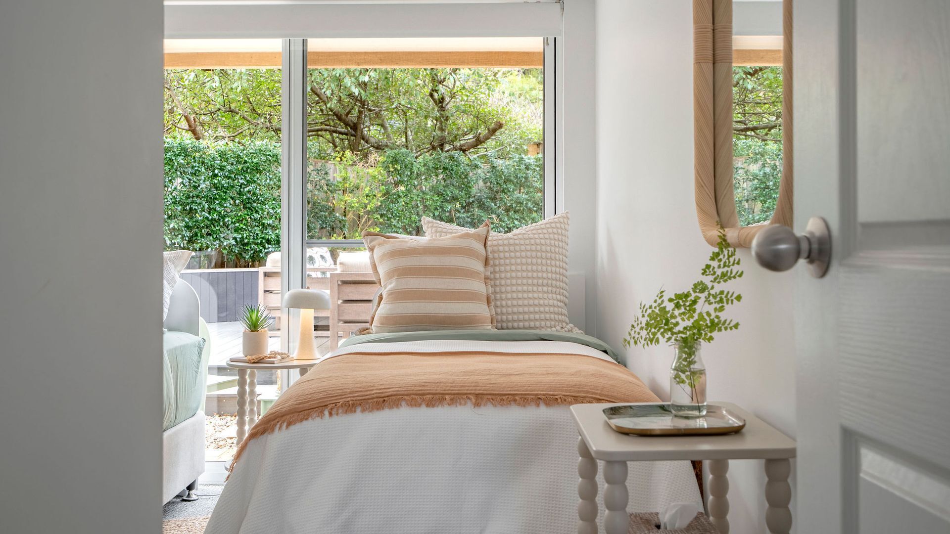 Ein Schlafzimmer mit einem Bett, Kissen, einem Nachttisch mit einer Vase und Blick auf Grünflächen.