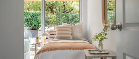 Ein Schlafzimmer mit einem Bett, Kissen, einem Nachttisch mit einer Vase und Blick auf Grünflächen.