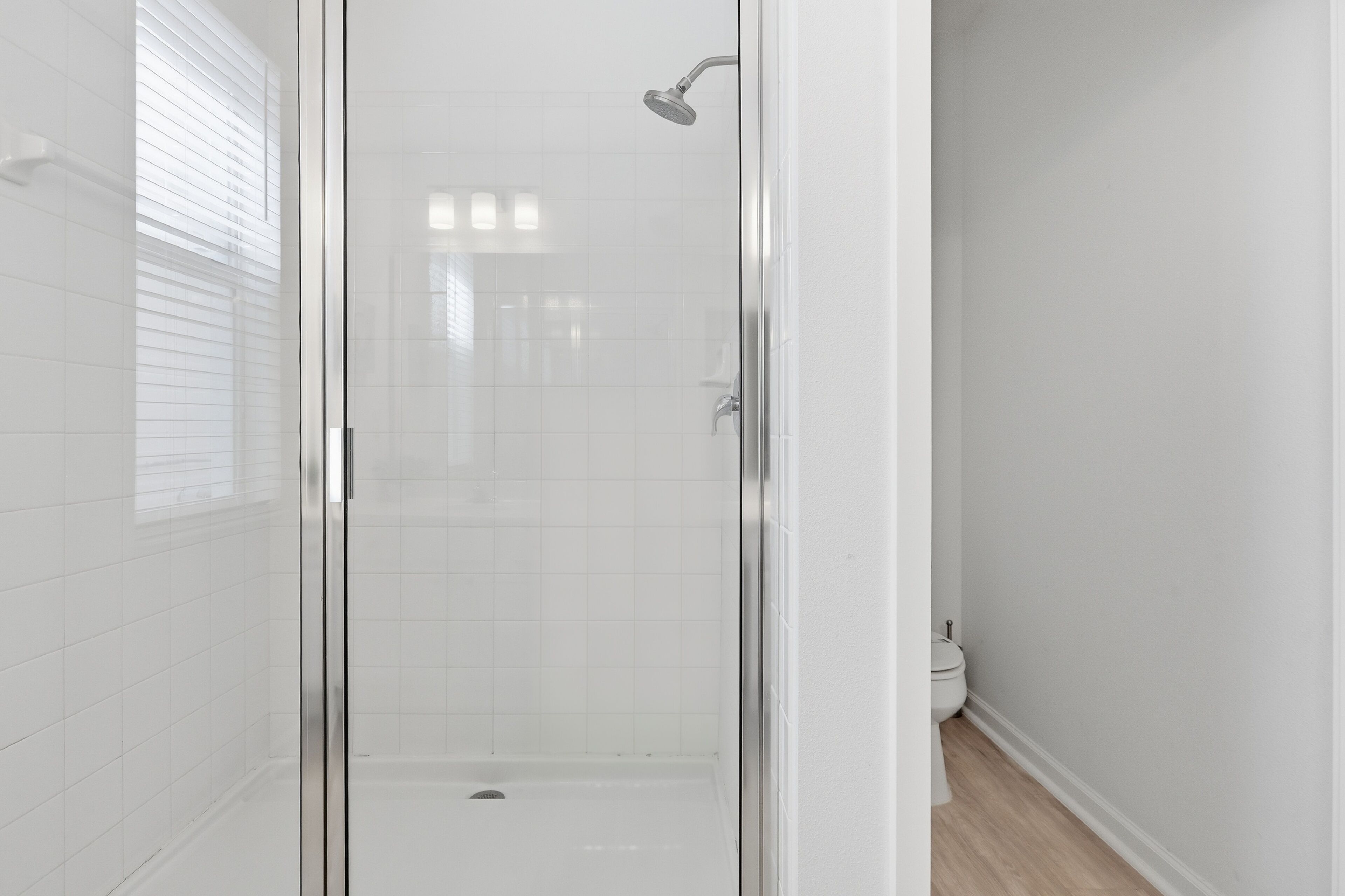 Une douche fermée par des parois de verre, dotée d’un pommeau de douche à main, avec des murs recouverts de carreaux blancs et des toilettes visibles dans la pièce adjacente.