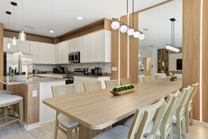 A modern kitchen with white cabinets, a central island, and a dining area with a long wooden table and chairs. - Disney 6BR/4BA Themed Rooms & Pool LED TV BV4509 (Kissimmee)