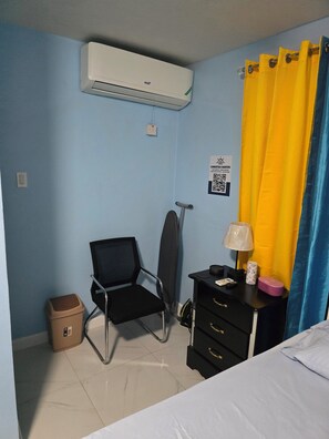 Habitación de hotel con una cama, un escritorio con lámpara, una silla, una tabla de planchar, un cubo de basura y un equipo de aire acondicionado.