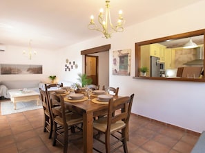 A dining area with a wooden table set for four, surrounded by chairs. A chandelier hangs above, and a large mirror reflects the kitchen area. - Portichuelo by Interhome (Almuñécar)