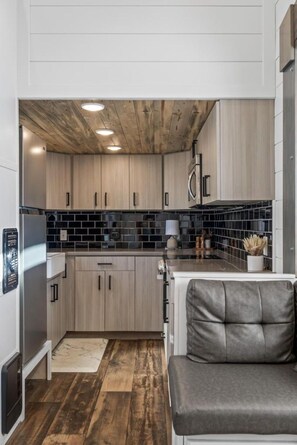 A compact kitchen with wooden cabinets, a black tiled backsplash, and a built-in microwave. - Modern Tiny Home | King Bed | A/C | Gas Fireplace (Donnelly)