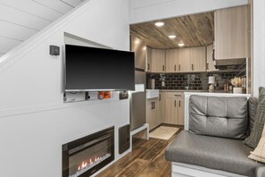 A modern interior with a flat-screen TV mounted on a white wall, a fireplace, a grey sofa, and a kitchen area with wooden cabinets and a tiled backsplash. - Modern Tiny Home | King Bed | A/C | Gas Fireplace (Donnelly)