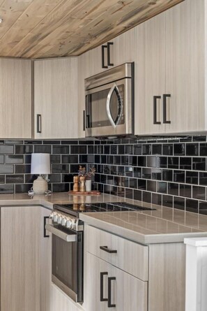 A modern kitchen with light wood cabinetry, a stainless steel microwave, and a black tiled backsplash. - Modern Tiny Home | King Bed | A/C | Gas Fireplace (Donnelly)