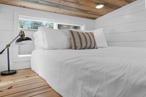 A bedroom with a bed, white bedding, a striped pillow, a desk lamp, and a window with blinds. - Modern Tiny Home | King Bed | A/C | Gas Fireplace (Donnelly)