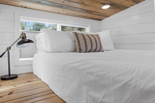 A bedroom with a bed, white bedding, a striped pillow, a desk lamp, and a window with blinds. - Modern Tiny Home | King Bed | A/C | Gas Fireplace (Donnelly)