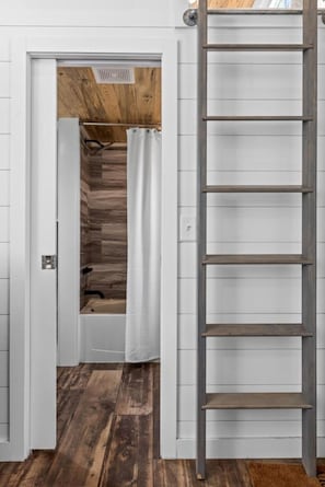 A bathroom with a shower, a wooden floor, and a white door. - Modern Tiny Home | King Bed | A/C | Gas Fireplace (Donnelly)