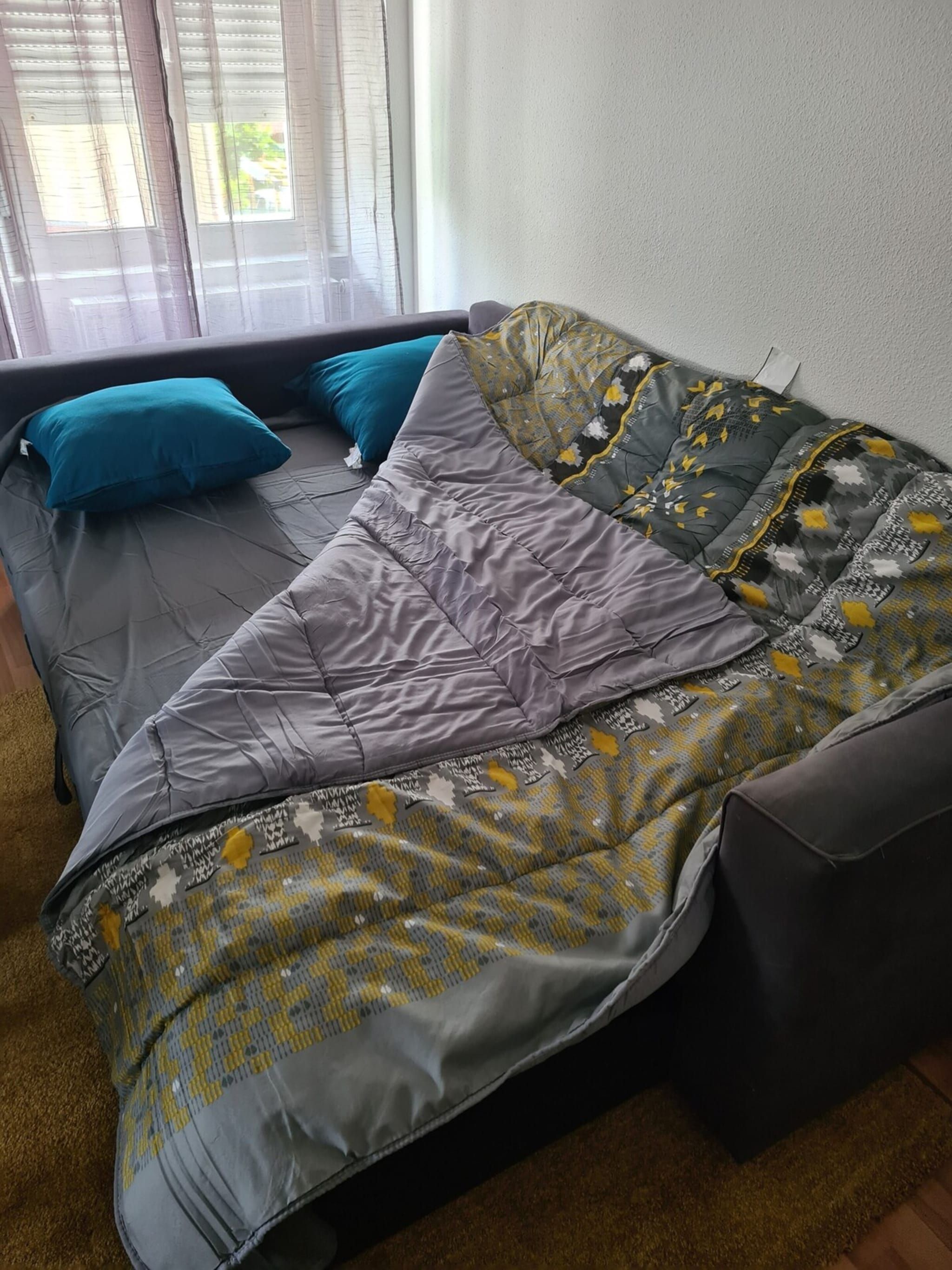Ein Bett mit grauem Matratze, Kissen in Türkis und Grau sowie einer gemusterten Decke.